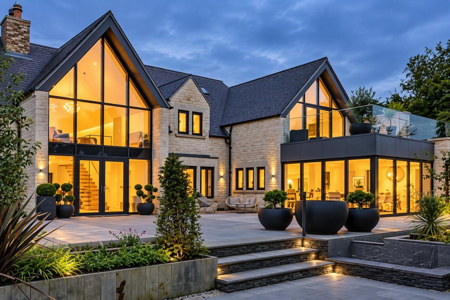 Stone house with large glazed gables and a lit garden terrace at dusk